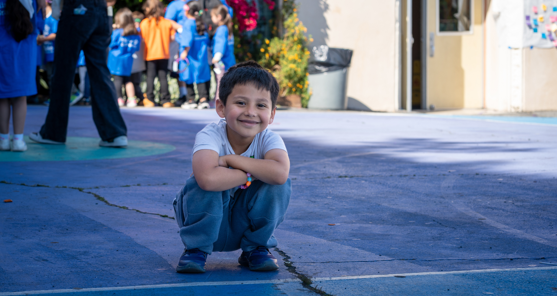 Smiling boy in gray shirt squats on a blue playground surface while a group of children in blue shirts stands in the background.