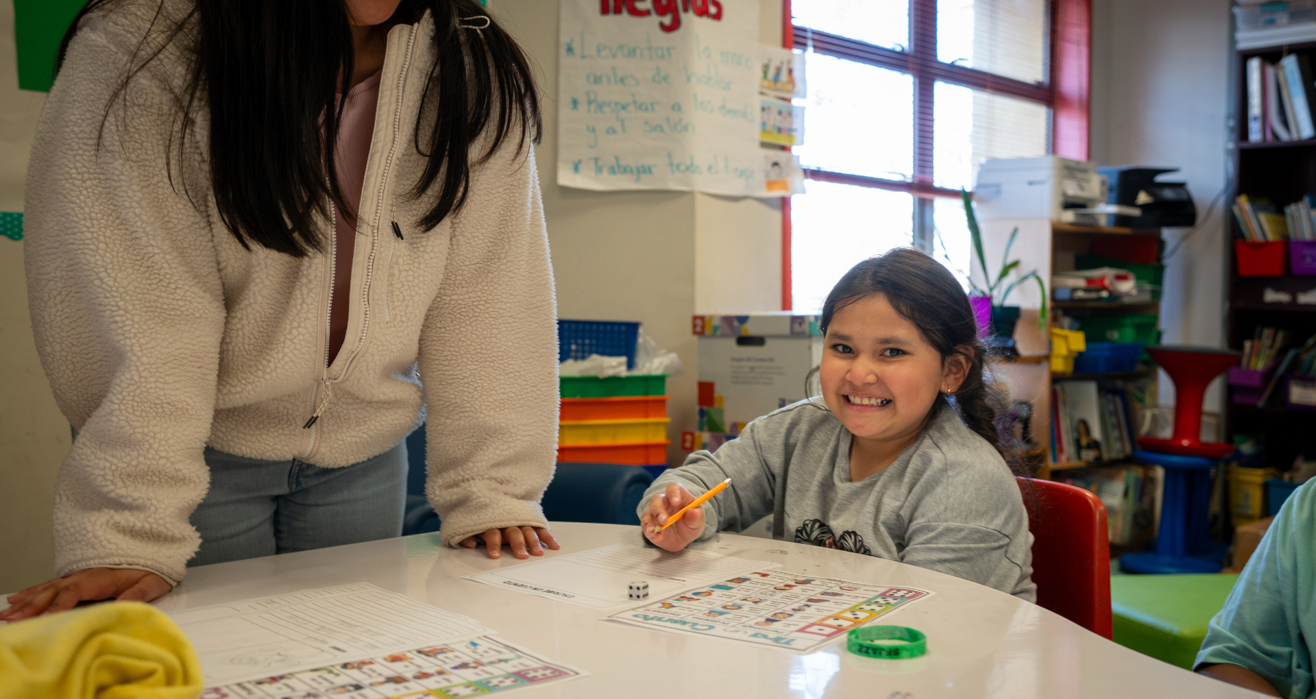 Young girl smiles while drawing at a table in a colorful classroom, with an adult leaning over nearby.