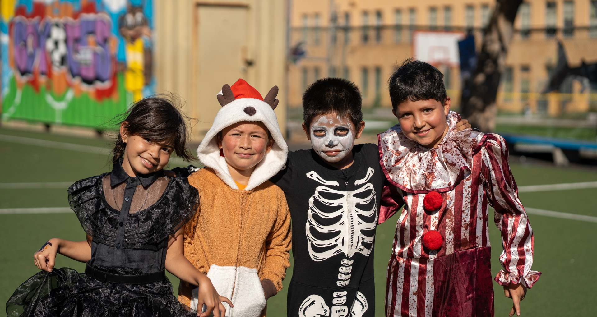 Four children in Halloween costumes pose together on a sunny schoolyard field, smiling at the camera.